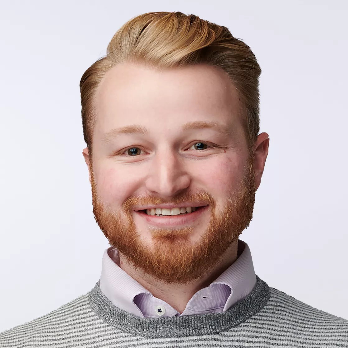 A person wearing a light gray striped sweater layered over a light pink collared shirt. The background is plain white, emphasizing the clothing and hairstyle. The image conveys a clean and professional appearance.